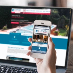 A hand holds a mobile phone displaying the Cornish Hot Tubs website, whilst a laptop in the background shows the same website with an image of a garden hot tub.